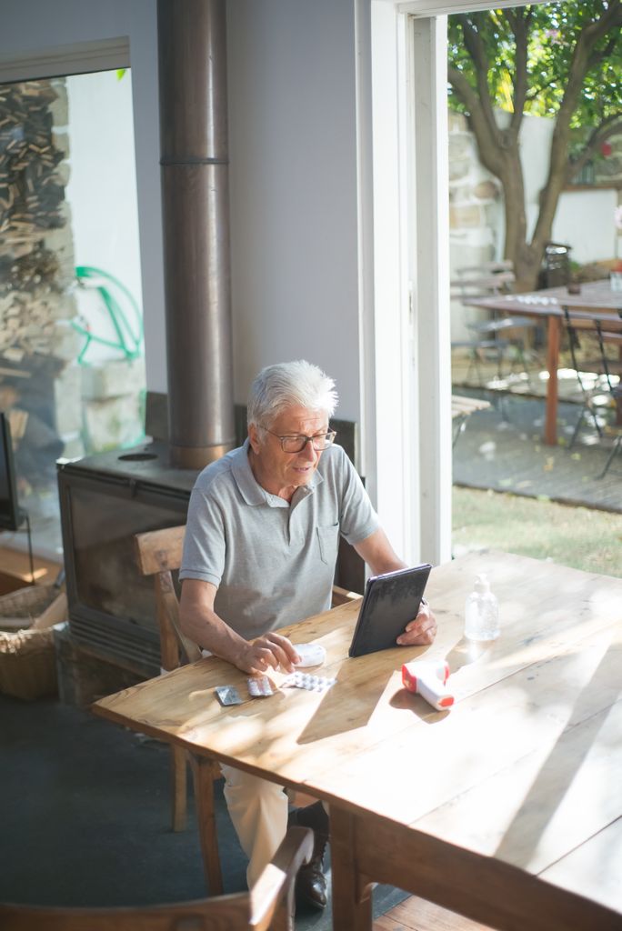 Elderly man with glasses reading a tablet at home, seated at a table with medicines, surrounded by natural light.